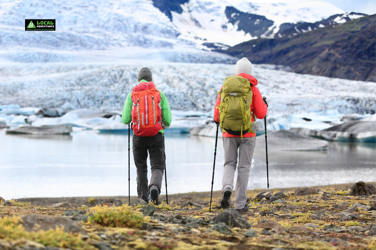 Two hikers in colorful jackets and backpacks walking with trekking poles toward a glacier in Iceland, showcasing the adventurous spirit of hiking in Iceland.