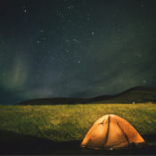 An orange tent set up under a starry night sky with faint northern lights in the background, showcasing Iceland tent camping in a serene landscape.