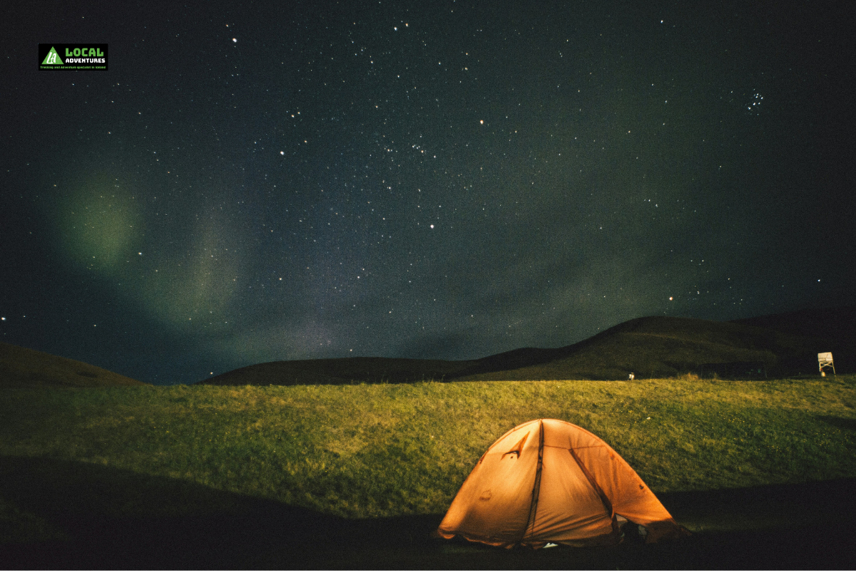 An orange tent set up under a starry night sky with faint northern lights in the background, showcasing Iceland tent camping in a serene landscape.