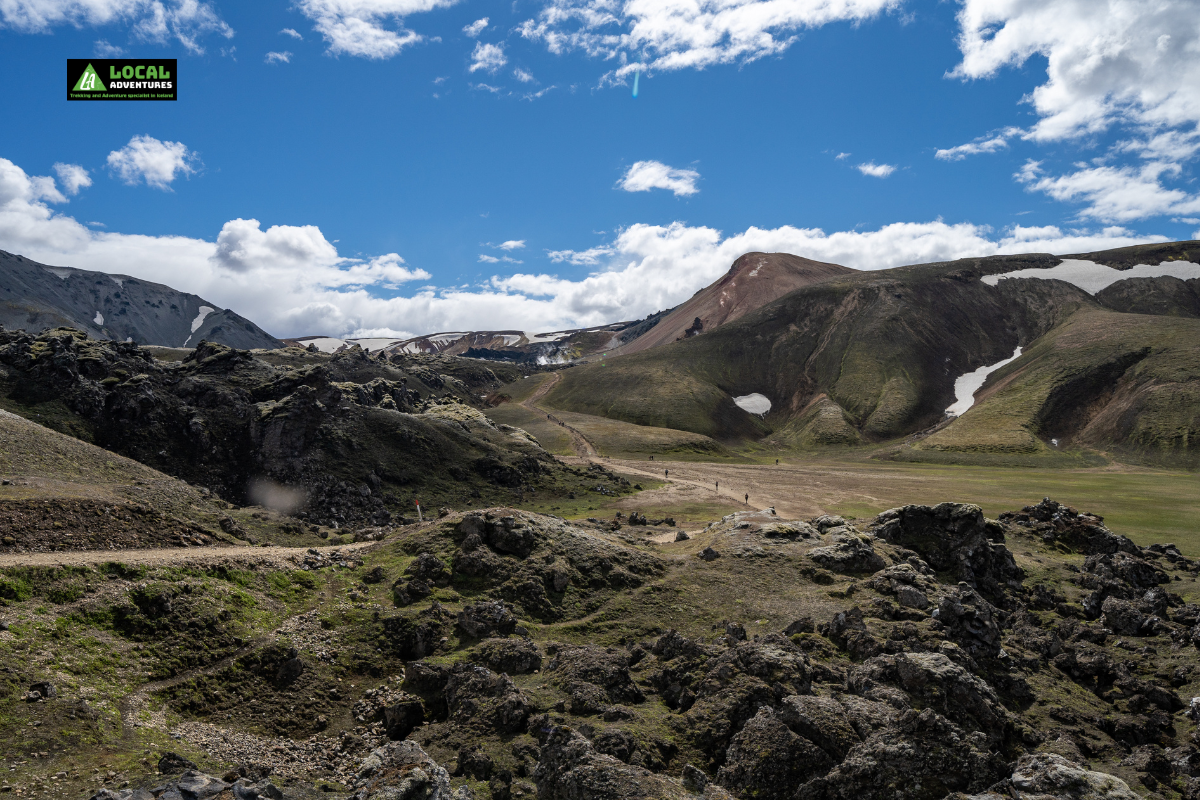 A scenic view of the Laugahraun Lava Field in Iceland, featuring rugged, moss-covered lava formations in the foreground with colorful mountains and patches of snow in the background. The sky is bright and partly cloudy, adding contrast to the volcanic landscape. A "Local Adventures" logo is in the top left corner of the image.