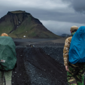 Two hikers wearing large backpacks walk along a rugged trail surrounded by volcanic terrain with green-covered mountains in the distance. The sky is overcast, creating a dramatic, moody atmosphere. A small logo for "Local Adventures" is in the top left corner of the image.