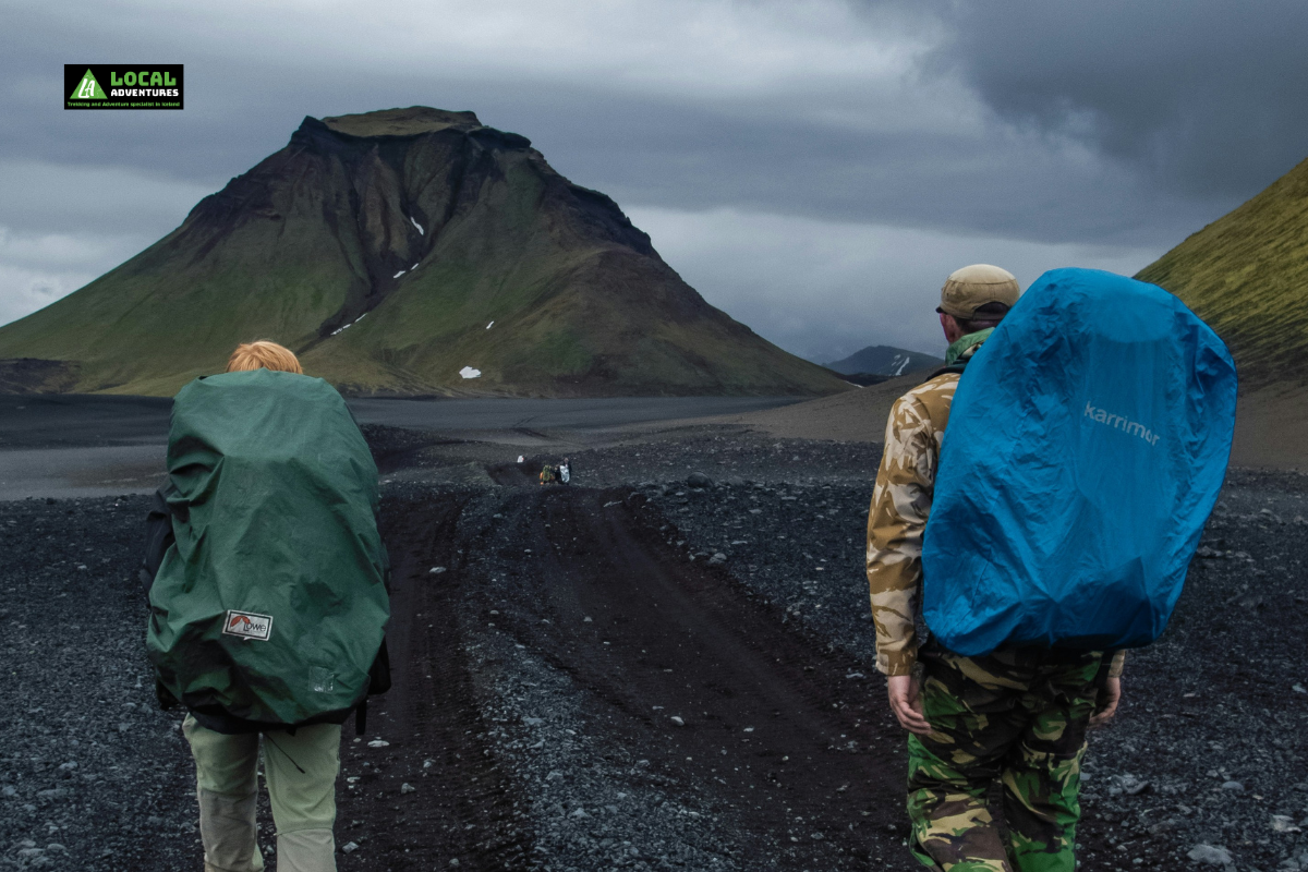 Two hikers wearing large backpacks walk along a rugged trail surrounded by volcanic terrain with green-covered mountains in the distance. The sky is overcast, creating a dramatic, moody atmosphere. A small logo for "Local Adventures" is in the top left corner of the image.