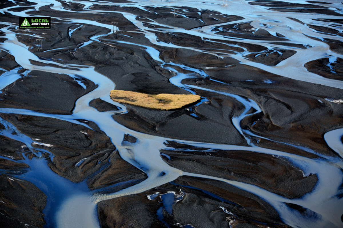 Aerial view of Markarfljót Canyon in Iceland, showcasing winding, braided rivers cutting through dark volcanic sands with a small, grassy island in the center. The intricate water channels create a striking contrast with the black landscape, forming a unique natural pattern. A "Local Adventures" logo is in the top left corner of the image.