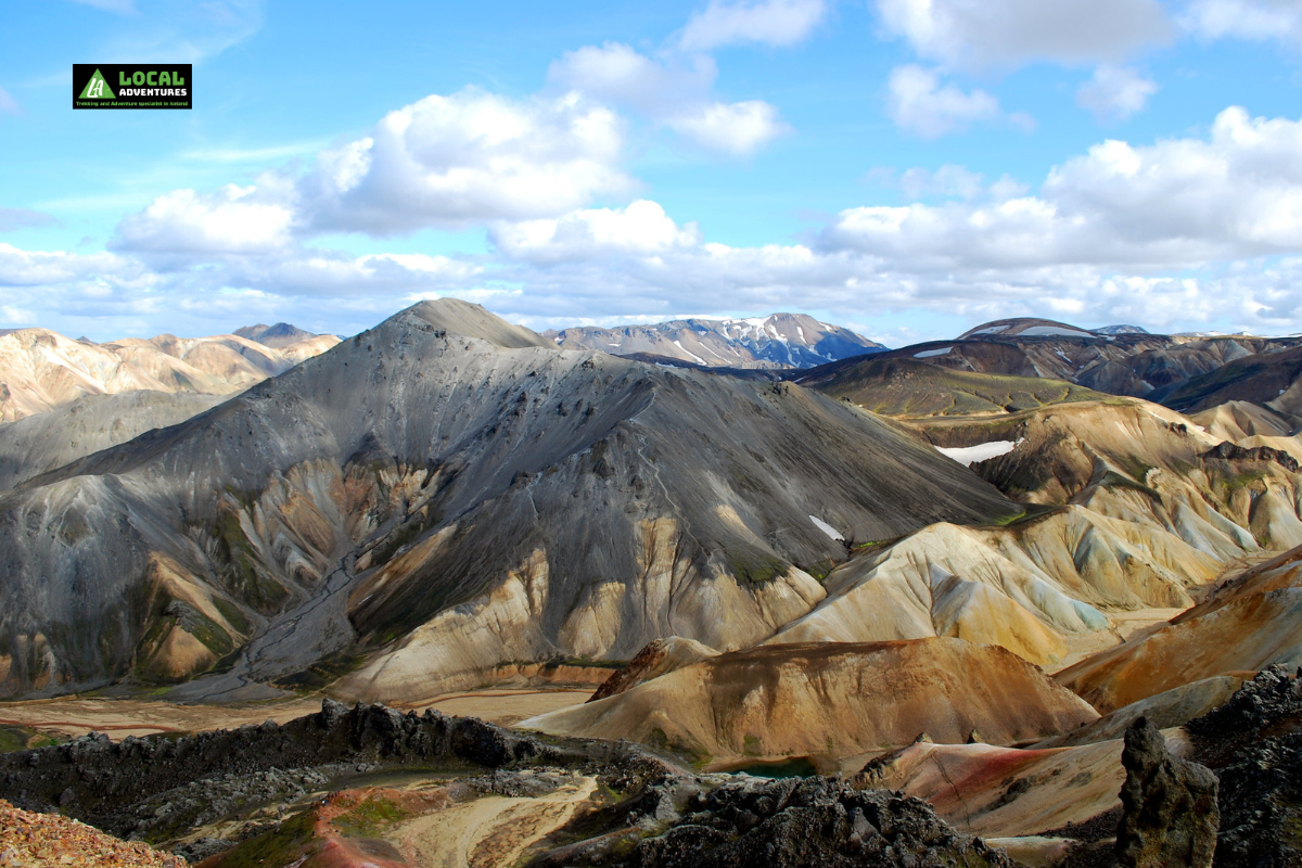 A breathtaking view of Mt. Bláhnúkur, also known as The Blue Peak, in Iceland. The mountain's unique gray and blueish tones stand out against the vibrant colors of the surrounding rhyolite hills. The scene is illuminated by sunlight under a partly cloudy sky, highlighting the rugged, layered texture of the volcanic landscape. A "Local Adventures" logo is in the top left corner of the image.