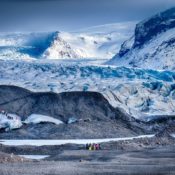 A group of hikers in colorful jackets walks across the expansive, icy terrain of Skaftafell Glacier in Iceland, with rugged mountain peaks and snow-covered landscapes in the background.