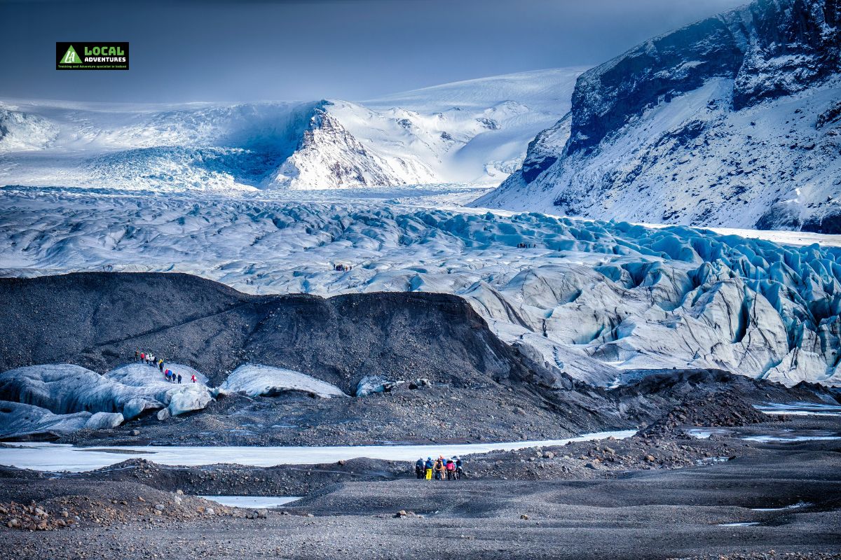 A group of hikers in colorful jackets walks across the expansive, icy terrain of Skaftafell Glacier in Iceland, with rugged mountain peaks and snow-covered landscapes in the background.