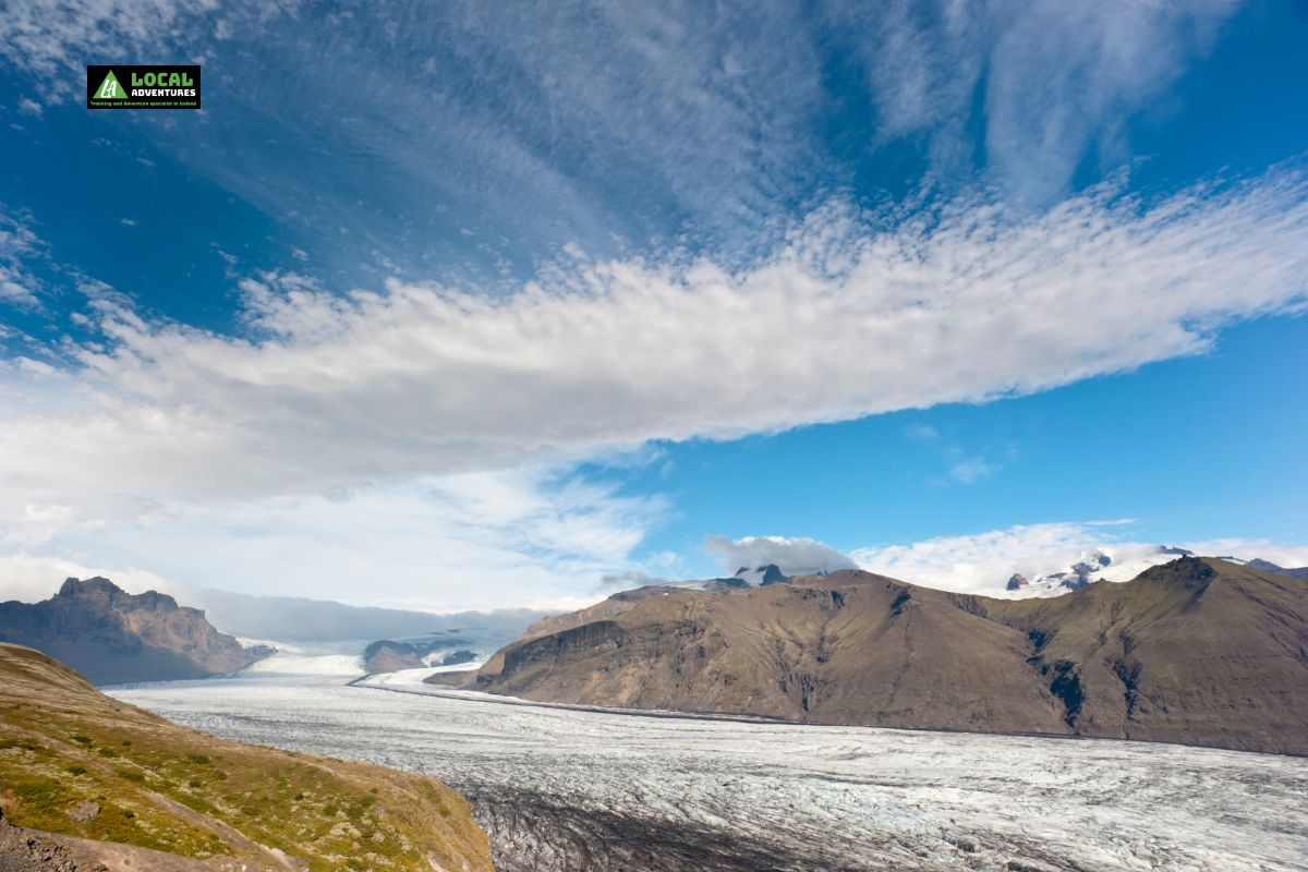 A scenic view of Skaftafell Glacier stretching across a valley, with rugged mountains and a bright blue sky filled with wispy clouds in the background.