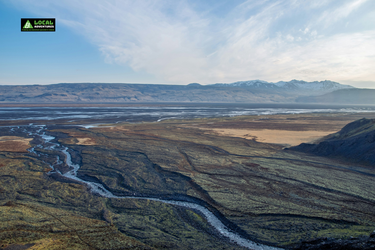 A panoramic view of Þórsmörk, also known as the Valley of Thor, in Iceland. The landscape features vast, moss-covered plains with a winding river cutting through, framed by distant snow-capped mountains under a clear sky. The expansive valley captures the rugged and serene beauty of Iceland's highlands. A "Local Adventures" logo is in the top left corner of the image.