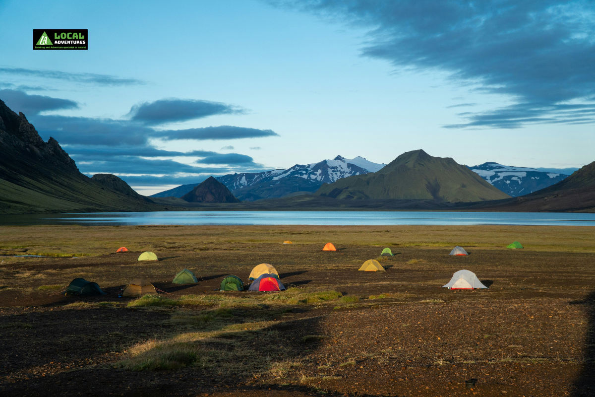 A scenic view of the Emstrur Valley in Iceland, showing a series of colorful tents scattered across an open, grassy plain near a calm lake. Snow-capped mountains rise in the background under a partly cloudy sky, creating a serene and picturesque camping scene.